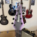 Detail hlavy elektrické kytary v fialové barvě s matným povrchem. Na hlavě je čitelné logo Ibanez a šest černých ladiček. V pozadí jsou vidět další k…