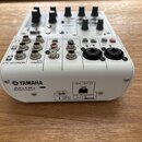 Bílá mixovací konzole s nápisem YAMAHA AG06 MIXING CONSOLE. Zadní strana obsahuje různé vstupy a výstupy, včetně portu USB 2.0 a konektorů pro napáje…