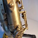 Detail saxofonu v matném zlatém provedení. Na korpusu je vyryto logo ve tvaru stylizovaného ptáka a pod ním nápis N.714155. Povrch nástroje vykazuje…