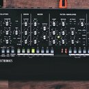 Černý analogový synthesizer s nápisy SE-02 STUDIO ELECTRONICS a logo Roland. Přední panel obsahuje množství otočných knoflíků, přepínačů a řadu bílýc…