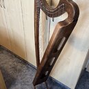 Tmavě hnědá dřevěná harfa s kovovými strunami. Nástroj má vyřezávaný rám s třemi oválnými otvory v bočním sloupci. Povrch dřeva je lakovaný a vykazuj…