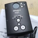 Černý ovládací modul elektronických bicích s displejem a několika tlačítky. Na přední straně je bílé logo Roland a nápis V-Drums. Jsou zde viditelné…