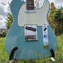 Světle modrá elektrická kytara typu Telecaster s bílým pickguardem a hnedým hmatníkem. Na snímači je viditelný nápis "Squier". Nástroj je umístěn na…