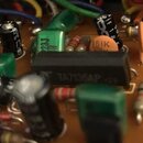 Detailní záběr na elektronickou desku s různými součástkami. Uprostřed je černý integrovaný obvod s bílým nápisem TA7136AP. Kolem jsou vidět kondenzá…