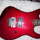 Detail elektrické kytary v červené barvě s kovovým hardwarem. Na mostku je viditelný nápis Floyd Rose. Kytara má jeden humbucker u mostku a hmatník s…