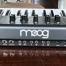 Černý syntetizátor s klaviaturou a řadou ovládacích knoflíků. Na přední straně je bílý nápis moog. Zadní panel obsahuje řadu konektorů s popisky: AUD…
