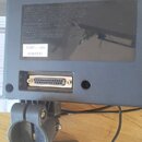 Šedý plastový box s montážní svorkou a připojeným napájecím adaptérem značky Roland. Na spodní straně boxu je vidět DB25 konektor a nálepka s textem…