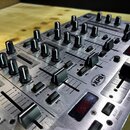 Stříbrný DJ mixér s kovovým povrchem a množstvím černých otočných knoflíků a posuvníků. Na panelu jsou viditelné nápisy „CH:2“, „LEVEL“, „LOW“, „EQ B…