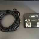 Černý pedál s nápisy LINE 6 a FBV2. Na povrchu jsou bílé šipky a ikona pěstí. K pedálu patří černý kabel s konektory RJ45.