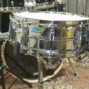 Stříbrný chromovaný snare drum s bílým blátcem. Na korpusu je modro-bílé logo Ludwig. Povrch chromu vykazuje drobné nečistoty a oprysky. Nástroj je u…