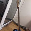 Elektrická kytara s modrým trupem a světlou dřevěnou hmatníkovou deskou. Na hlavě kytary nejsou žádné viditelné značky ani nápisy. Nástroj je opřen o…