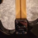 Detail krku a spoje s tělem kytary v černé barvě. Na chromovaném krytu spoje je vyryté logo Fender a nápis CORONA, CALIFORNIA. Povrch kytary i kovové…