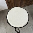 Fotografie zachycuje kulatý rámový buben s bílou blánou. Okraj nástroje je potažen černým materiálem a zdobený barevným provázkem. Z spodní části vyč…