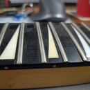 Detailní pohled na hmatník nástroje s černým povrchem a kovovými pražci. Mezi pražci jsou vloženy tři bílé trojúhelníkové markery. Povrch hmatníku vy…