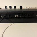 Detail zadní strany černého hudebního zařízení s texturovaným povrchem. Jsou vidět konektory a nápisy DC 12V, STANDBY a USB. V horní části jsou čtyři…