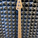 Fotografie zachycuje hmatník a hlavu elektrické basové kytary s bílým tělem. Na dřevěné hlavě je viditelné logo Fender a nápis JAZZ BASS. Hmatník je…