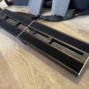Dvě černé obdélníkové podložky s vnitřními výřezy, vyložené černým pěnovým materiálem. Podložky mají stříbrné okraje a jsou umístěny na dřevěné podla…