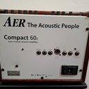 Zadní strana akustického zesilovače v hnědém dřevěném rámu s bílým předním panelem. Na panelu je nápis AER The Acoustic People® a model Compact 60₂ t…