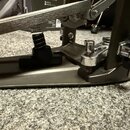 Detailní záběr na spodní část kovového pedálu v šedé barvě. Jsou vidět nastavitelné mechanismy s pružinou a chromovanými šrouby. Na povrchu jsou vidi…