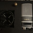 Kondenzátorový mikrofon stříbrné barvy s kovovou síťkou, umístěný v černé polstrované přepravce. Na těle mikrofonu je nápis „Vintage V67 S/N: 2334“ a…