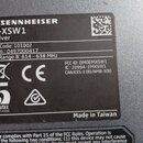 Fotografie detailně zachycuje identifikační štítek na černém povrchu zařízení. Na štítku je uveden název SENNHEISER, model EM-XSW1 Receiver, Prod. Co…