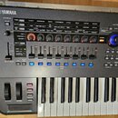 Šedý digitální syntezátor značky YAMAHA s klávesnicí a rozsáhlým ovládacím panelem. Panel obsahuje řadu posuvníků, otočných knoflíků a tlačítek s pop…
