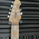 Fotografie zachycuje hlavu a část hmatníku elektrické kytary z přírodního dřeva. Na hlavě je černý nápis „Sterling by Music Man“ a nad ním černý podp…