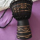Černý djembe s bílou membránou a černým provázkem. Tělo nástroje je zdobeno vyrytými světlými ornamenty v horní a dolní části. K nástroji je přivázan…