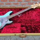Modrá elektrická basová kytara v otevřeném kufru s červeným sametovým vnitřkem. Na hlavě nástroje je viditelné logo Fender. Tělo kytary má stříbrný k…