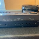 Zadní strana tmavého elektronického klavíru značky Roland. Jsou zde viditelné nápisy MEMORY PROTECT (ON OFF ON), TAPE (LOAD SAVE), EXT CONTROL (FOOT…