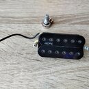 Černý humbucker s nápisem Bach v bílé barvě. K snímači je přiložen potenciometr s kovovým knoflíkem a nápisem BOURG na boku. Vše leží na světlém dřev…