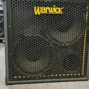 Černý basový reproduktor v šedém rámu s kovovou ochrannou mřížkou. V horní části je žluté logo Warwick. Za mřížkou jsou vidět dva velké reproduktory…