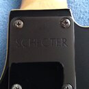 Detail krku a korpusu elektrické kytary v černé barvě. Na černém plastovém krytu krku je vyryt nápis SCHECTER. Kryt je připevněn čtyřmi křížovými šro…