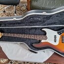 V otevřeném černém kufru s vnitřním čalouněním leží elektrická basová kytara. Nástroj má tělo v barvě sunburst, bílý pickguard a hnědý hmatník. Na hl…