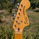 Fotografie zachicuje hlavu dřevěné elektrokytary v přirozené barvě. Na zadní straně jsou vidět nápisy Squier by Fender a STRATOCASTER. Kytara má šest…