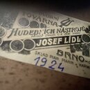 Fotografie detailu papírové štítku s nápisy. Je na něm uveden text „DVORNÍ TOVÁRNA HUDEBNÍCH NÁSTROJŮ“, „PRISEZ. ZNALEC C.K. ZEM. SOUDU.“ a jméno „JO…
