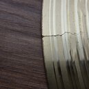 Detailní záběr na zlatý kovový povrch s vertikálním žebrováním, který je umístěn vedle tmavého dřeva. V kovové části je viditelná vodorovná prasklina…