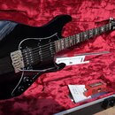 Černá elektrická kytara s tvarem typu Stratocaster v otevřeném kufru s červeným vnitřkem. Na hlavě kytary je bílý nápis Ibanez. Kytara má chromovaný…