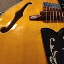 Detailní pohled na tělo žluté polodupaté elektrické kytary s f-otvorem. Na dřevěném kobylníku je viditelné logo znacky Epiphone. Kytara má dva chromo…
