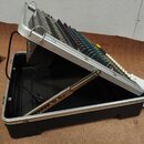 Černý přenosný kufr s hliníkovým profilem, v němž je umístěn elektronický nástroj s řadou barevných tlačítek a přepínačů. Z přístroje vede černý napá…