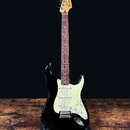 Černá elektrická kytara s bílým pickguardem a hnedým hmatníkem. Na hlavě je viditelné logo Fender Stratocaster. Nástroj má tři snímače typu single-co…