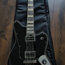 Černá elektrická kytara s hnědým hрифem a bílými vložkami na hmatníku, položená na černém obalu. Na hlavě je viditelné logo "Gretsch". Kytara má dva…