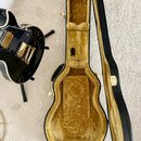 Černá elektrická kytara s gold hardwarem a černým korpusem, umístěná na stojanu. Vedle ní je otevřený černý tvrdý kufřík s žlutým plyšovým vnitřkem.…