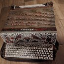 Akordeon s červeným, modrým a zlatým ornamentálním vzorem na mechu a korpusu. Na horní části je bílý nápis KEBROLE. Nástroj má bílé klávesy v horní č…