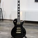 Černá elektrická kytara s gold hardwarem umístěná na černém stojanu. Na hlavě hředu je bílý nápis Epiphone a ozdobný vzor. Kytara má dva humbuckery,…