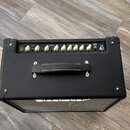Černý kytarový kombo zesilovač s koženým povrchem a bílými ovládacími knoflíky. Na boku je vidět část nápisu BLOKER. Ovládací panel obsahuje popisky…