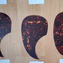 Tři kytarové plektra v podobě velkých krytek (pickguardů) s tmavě hnědou a žlutou mramorovou texturou. Jsou vyrobeny z lesklého materiálu a mají zakř…