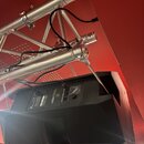 Černý stage monitor s hranatým tvarem, umístěný pod hliníkovou konstrukcí z trubek. Na zadním panelu je viditelný nápis "POWERBOOST" a logo ve tvaru…