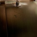 Detailní záběr na korpus bubnu v tmavě zelené barvě s kovovým lugem a šroubem. Povrch nástroje vykazuje viditelné opotřebení ve formě škrábanců a odř…