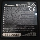 Fotografie detailu černé štítku s bílým textem. V horní části je logo Ibanez a nápis AC ADAPTER DC9V s grafickým znázorněním konektoru a značka CE. D…